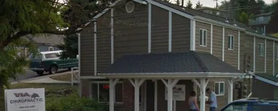 Two-story dark wooden house with covered front porch and white trim columns, parked vehicles visible on residential street