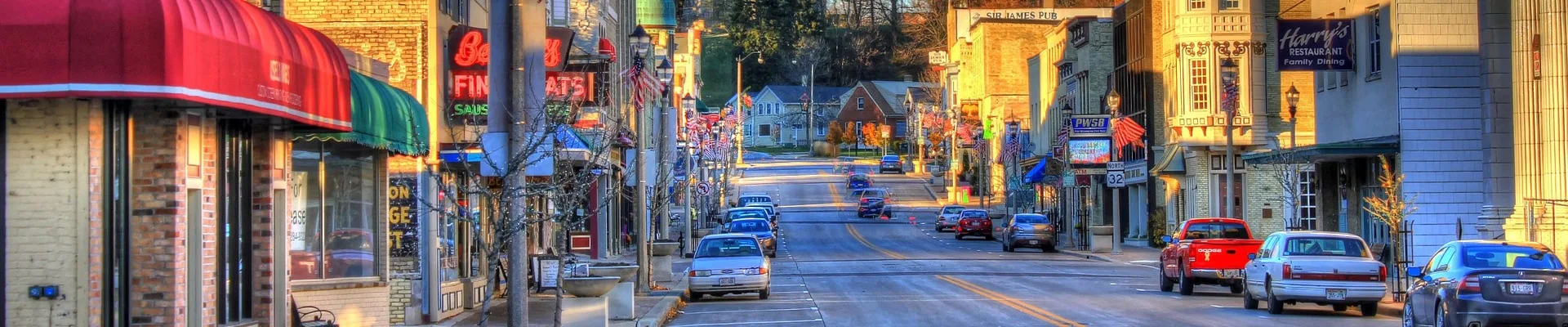 Downtown street at dusk with storefronts, neon signs, and vintage cars parked along the road